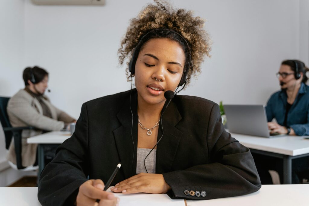 African American woman with headset working in an office with colleagues, using a pen for note-taking.